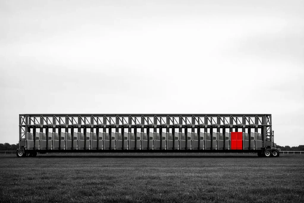 A row of empty racecourse starting stalls on dark turf, the gate of one stall painted red