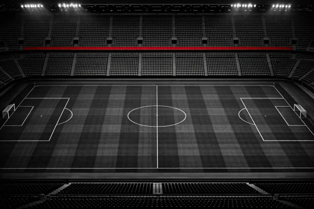 Empty floodlit football stadium seen from the upper tier at night, the pitch markings picked out against dark turf.