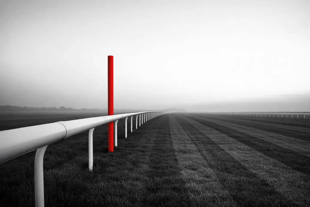 The white running rail of an empty racecourse home straight at dawn, one red marker pole standing beside it